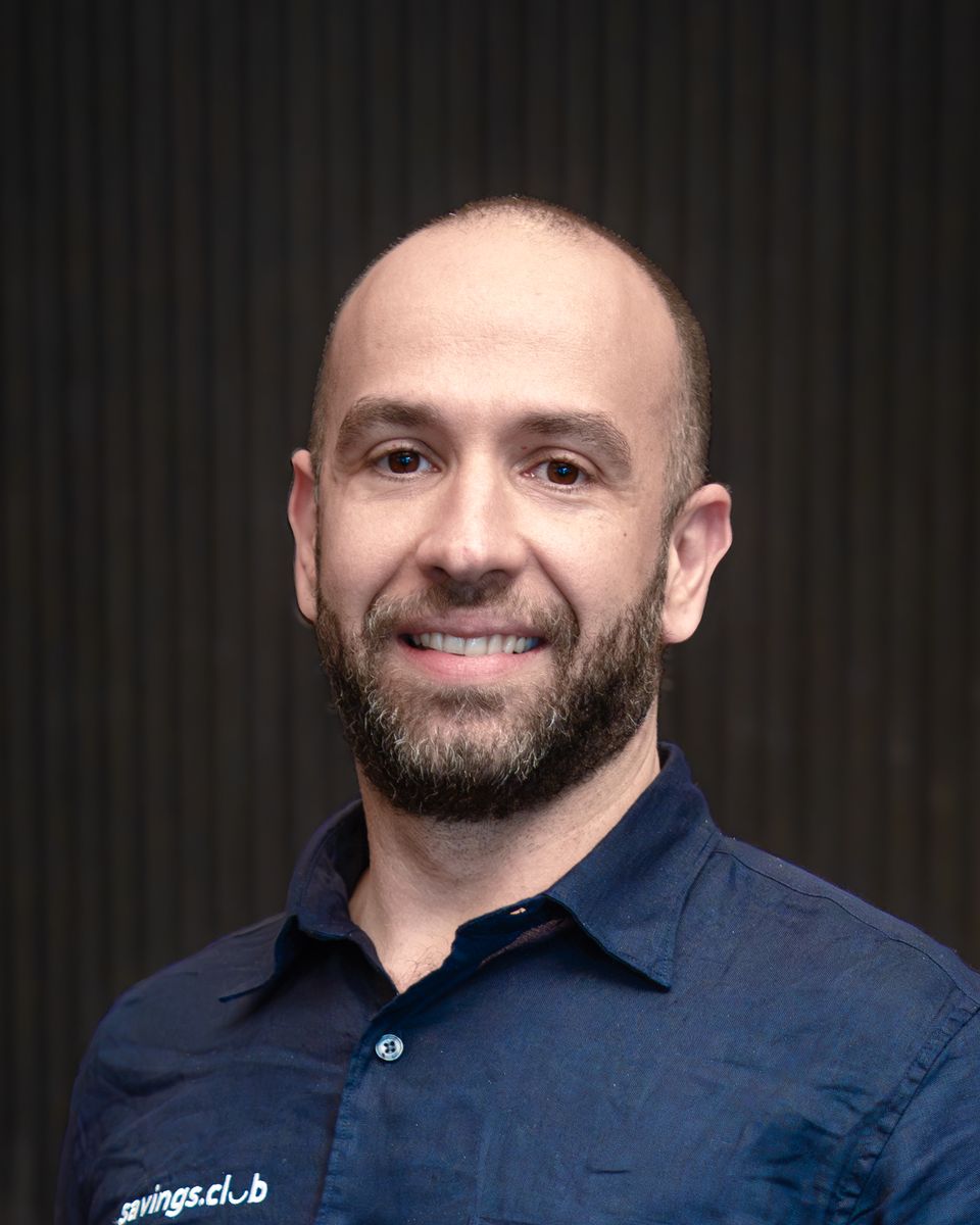 Adriano Marques in a navy shirt with the savings.club logo, in front of a dark slatted wall.